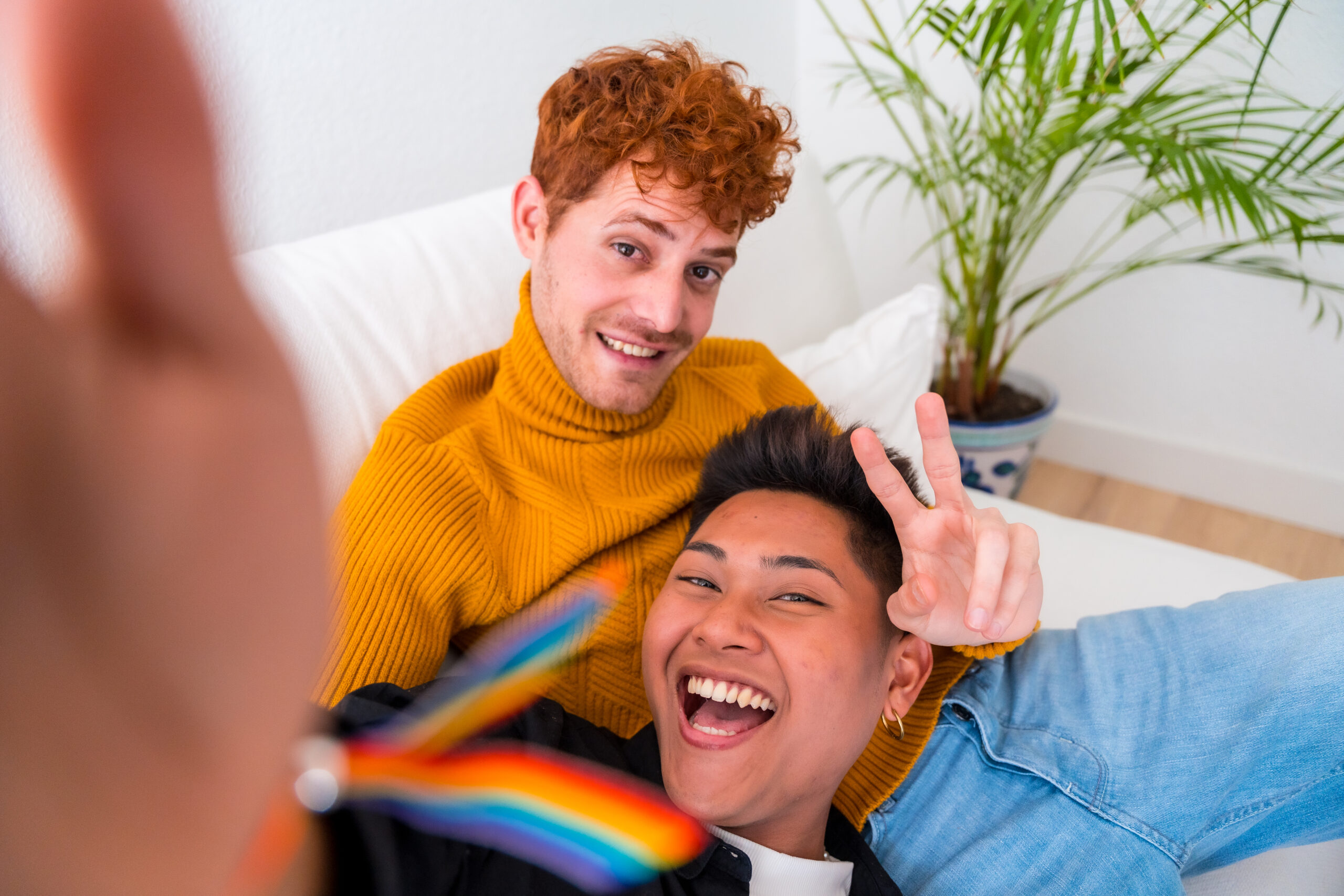 Beautiful gay couple being romantic indoors at home on the sofa, gay couple taking selfie and making victory sign, lgbt concept