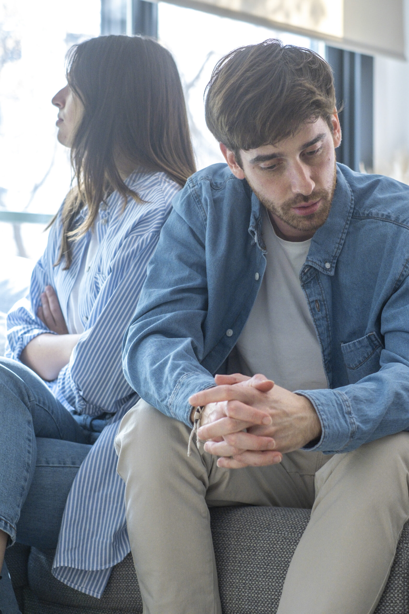 Unhappy couple sitting on sofa at home looking to opposite directions sad. Young adult man and woman upset and concerned. People with relationship problems to solve living together in apartment